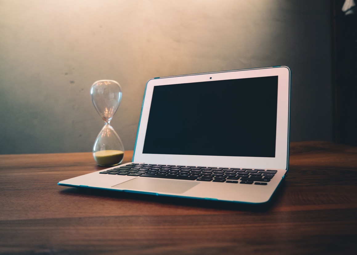 An hourglass beside an open laptop on a wooden desk