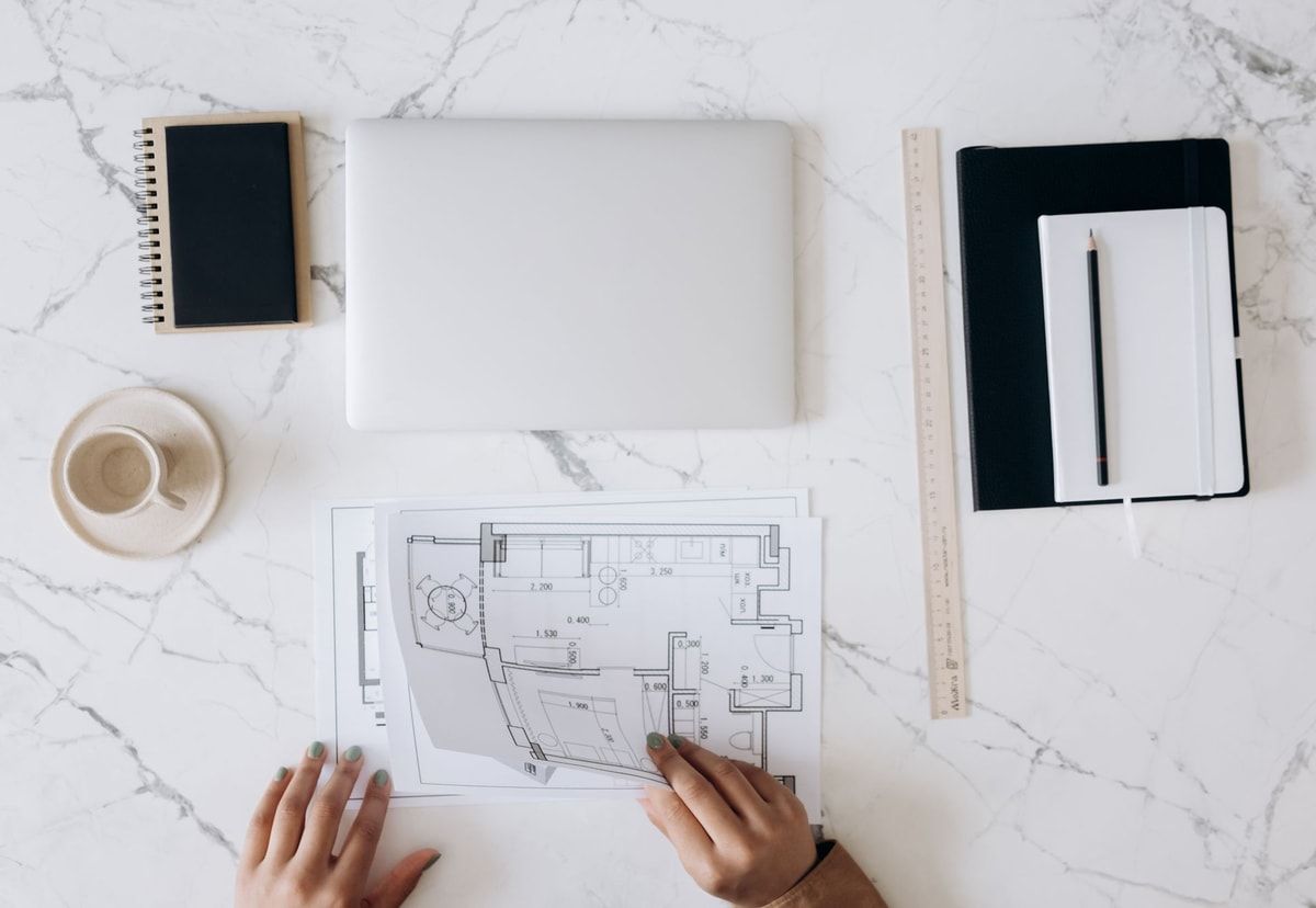 Top-down view of hands reviewing architectural floor plans beside a laptop, ruler, notebooks and coffee on a light desk